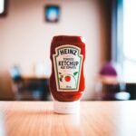 A Heinz Tomato Ketchup bottle on a wooden table with blurred background indoors.